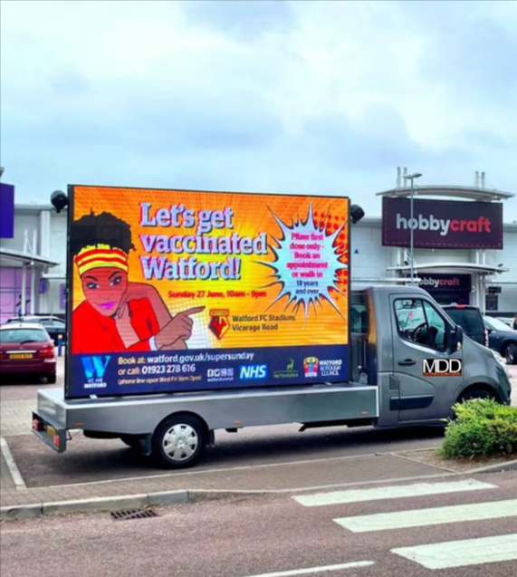 Mobile Digital Displays MDD DigiVan with large LED screen showing NHS vaccination campaign advertisement in UK retail park car park